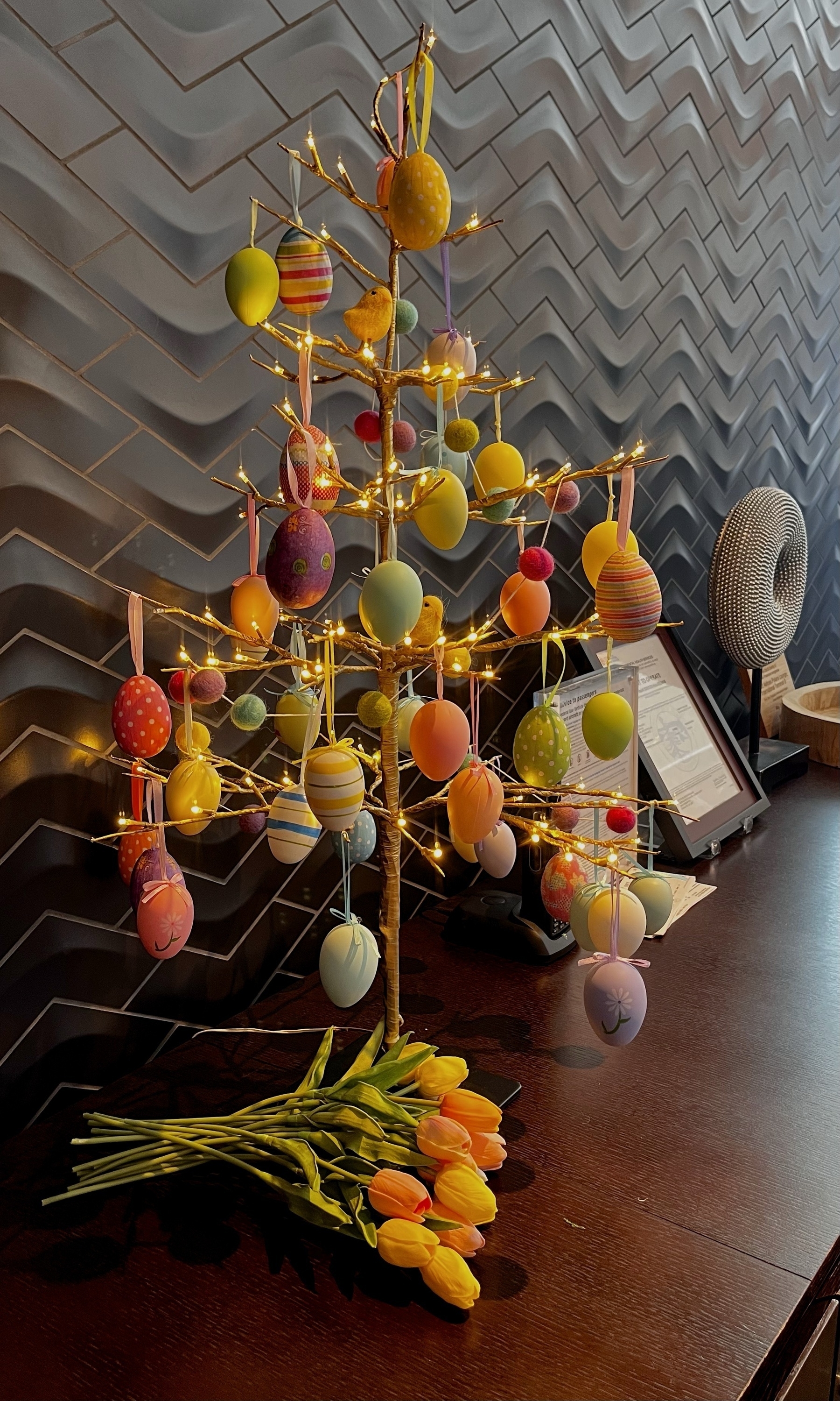 A small decorative tree decorated with small Easter eggs and fairy lights. Some peach colored tulips lie at its base