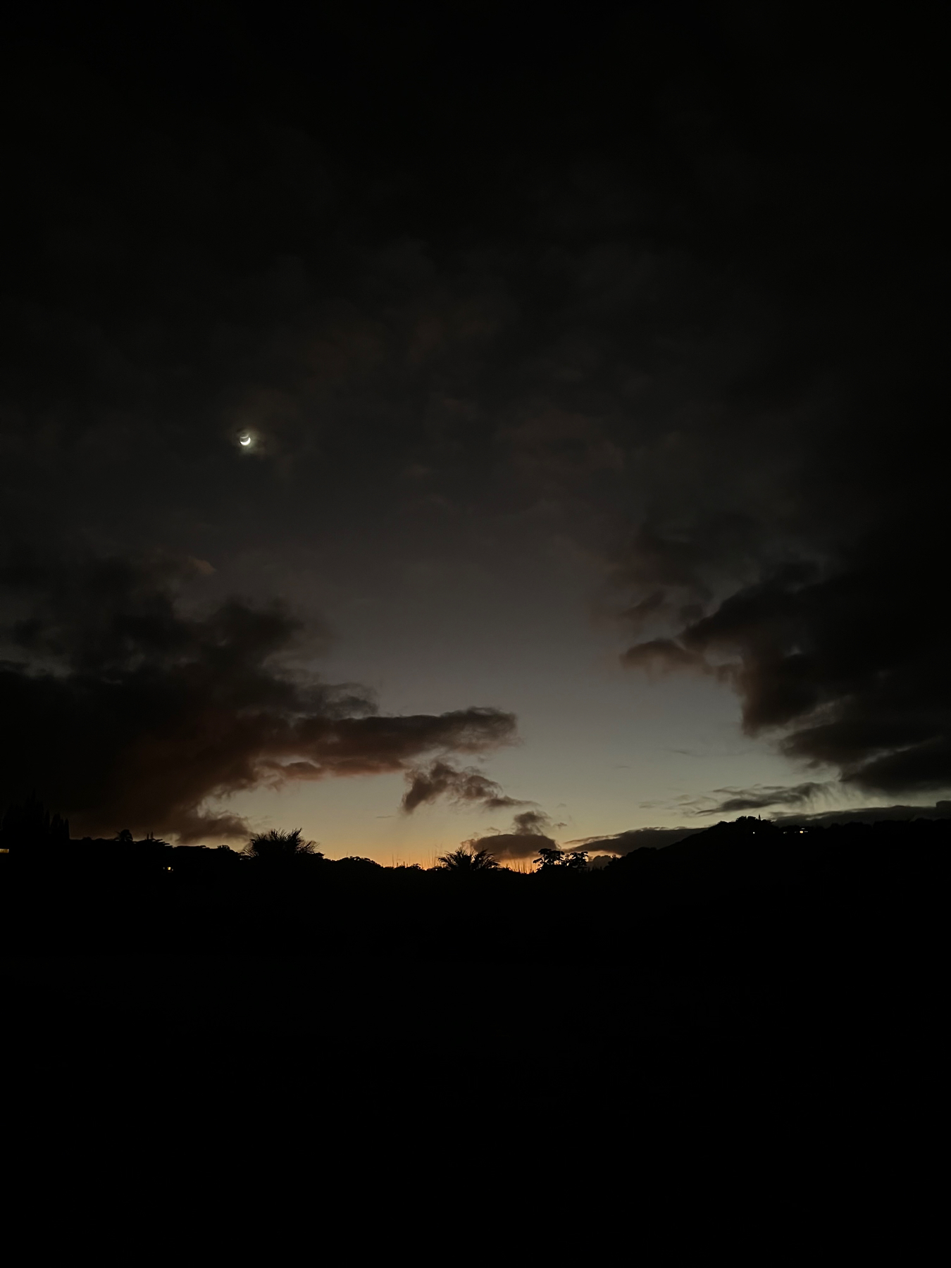 A dark sunset sky with clouds and a crescent moon, with silhouettes of trees and hills on the horizon.
