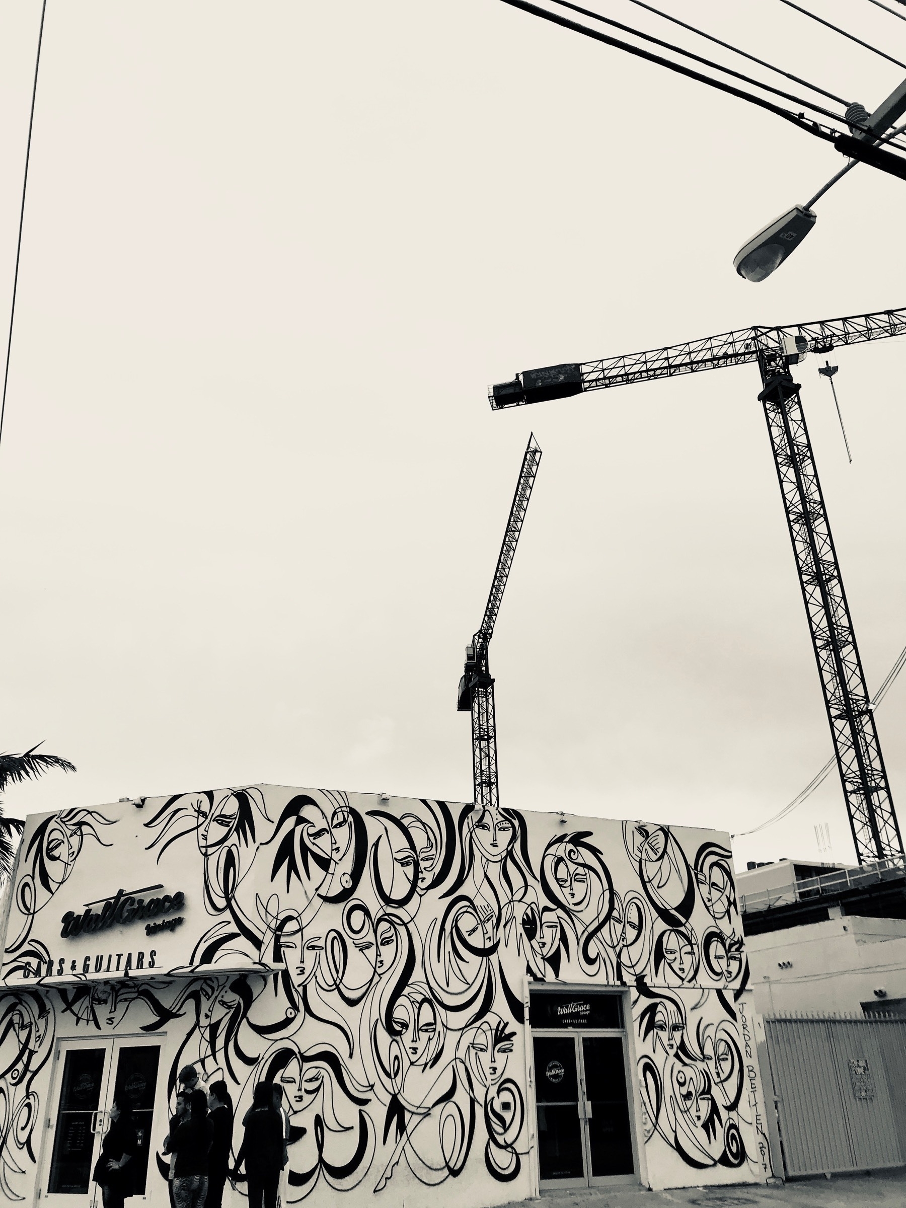 A black and white photograph of cranes appearing above a single story building with an abstract mural painted on it
