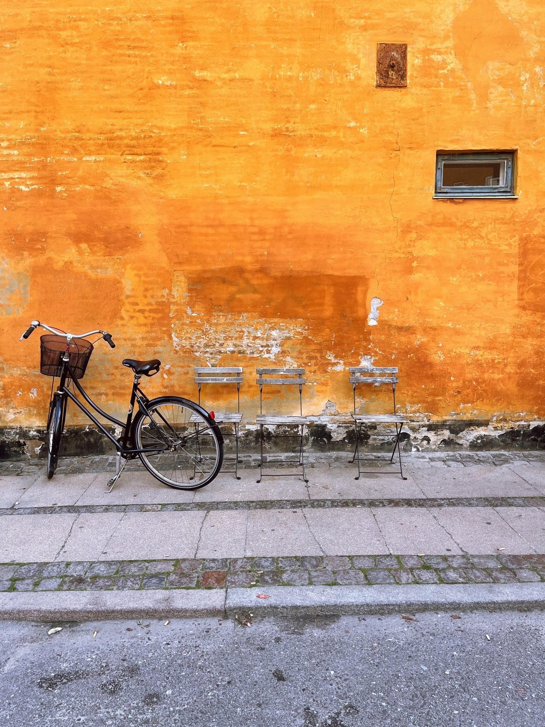 A bicycle and three chairs next to an orange wall, on a street in Copenhagen