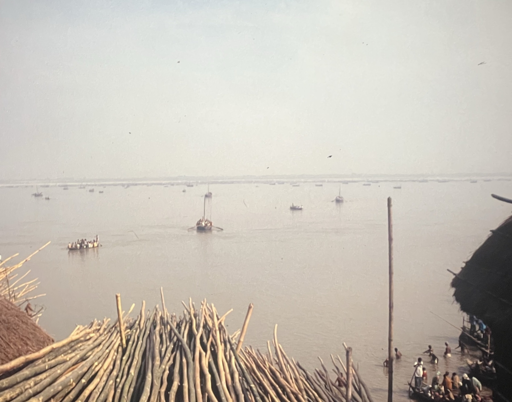 Looking down on a view over the River Ganges. It is a hazy view. In the foreground, upon which I might be sitting, are a pile of wooden poles, a couple of thatched roofs are to each side. The river has a variety of boats on them of different sizes, all full of people, and none motorized but rowed