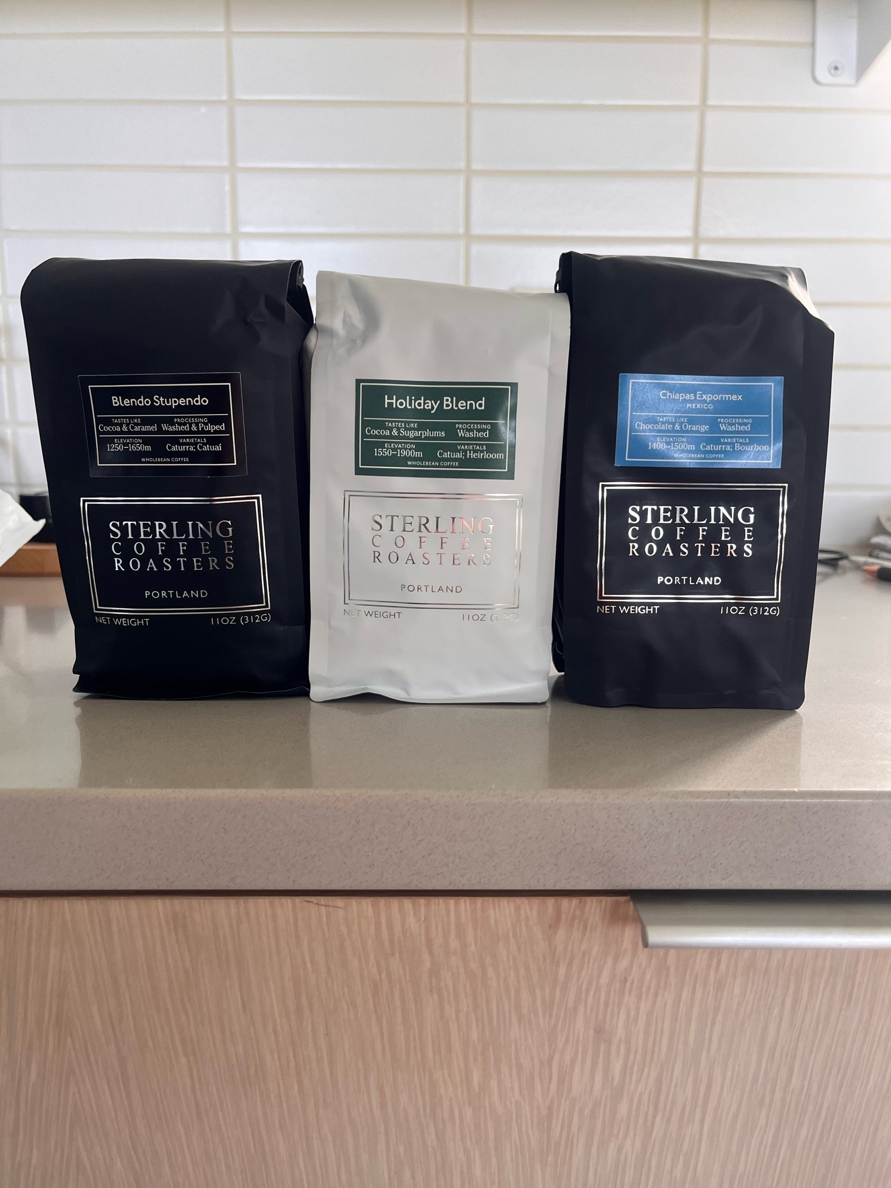 Three bags of Sterling Coffee Roasters coffee on a kitchen counter: “Blendo Stupendo,” “Holiday Blend,” and “Chiapas Eupromex.” The bags are labeled with flavor notes, processing methods, and elevation of the coffee beans