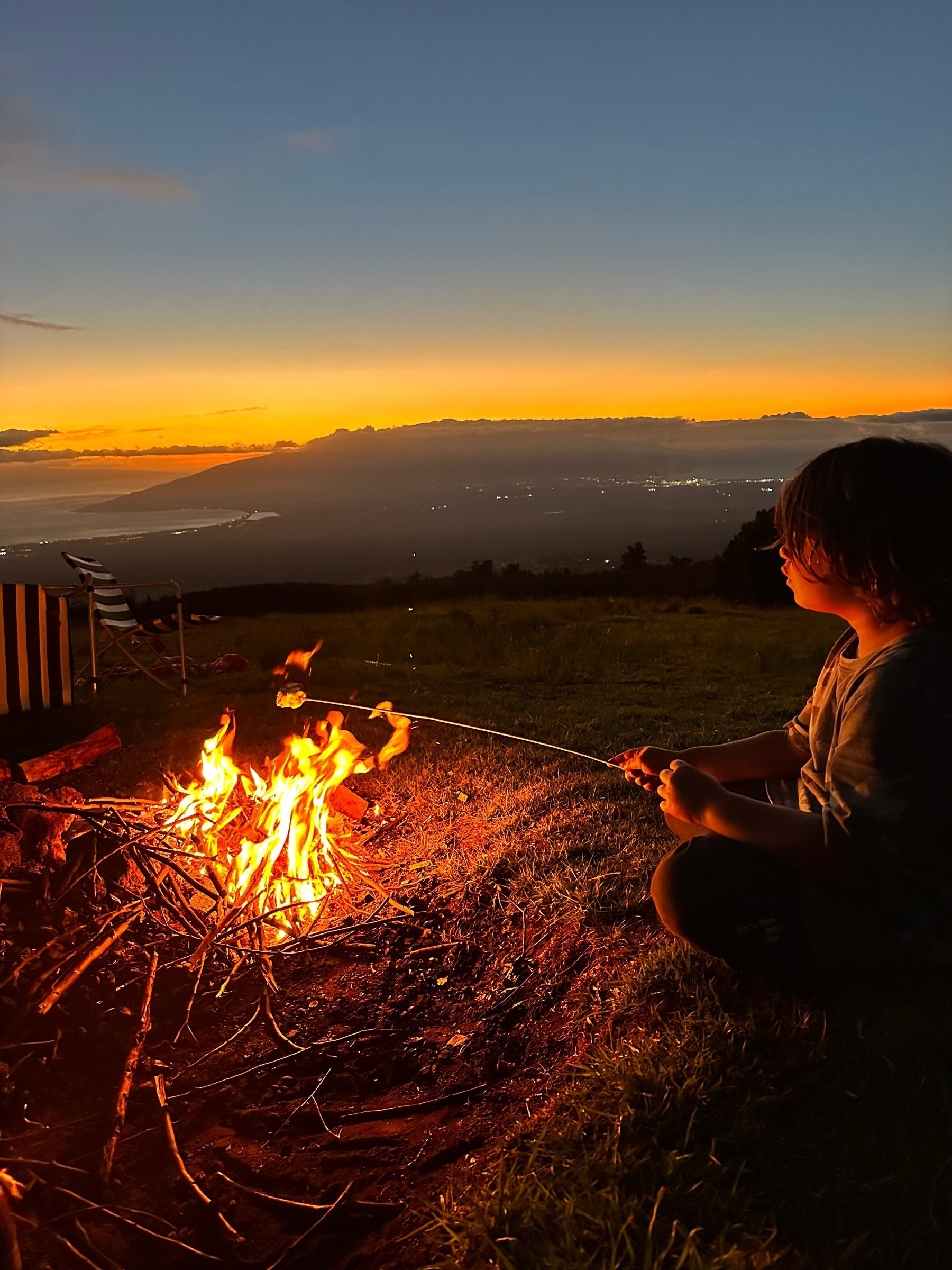 A boy roasting a marshmallow over an open fire at dusk in PoliPoli Springs Recreation Area, Maui