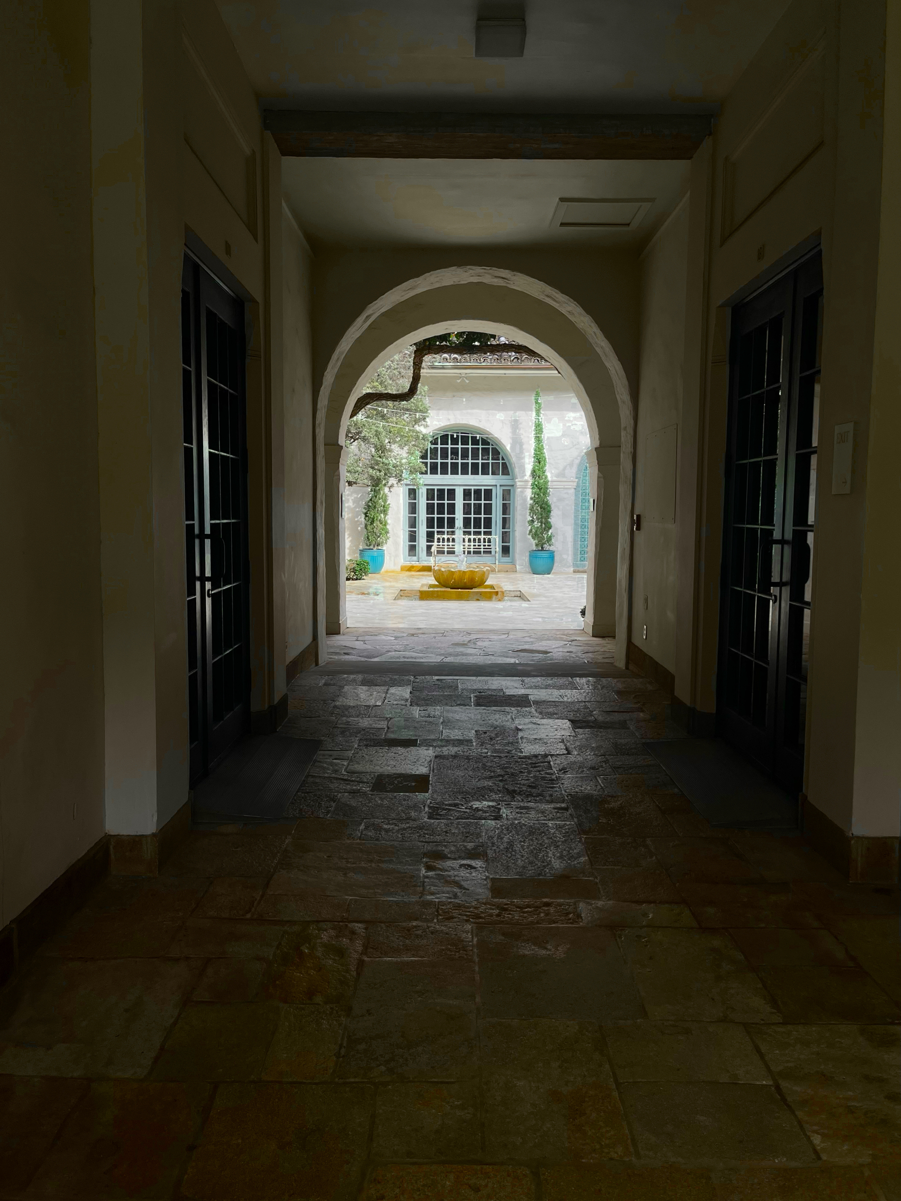 A hallway with stone flooring, wooden doors, and aged walls, leads to a bright courtyard with a fountain.
