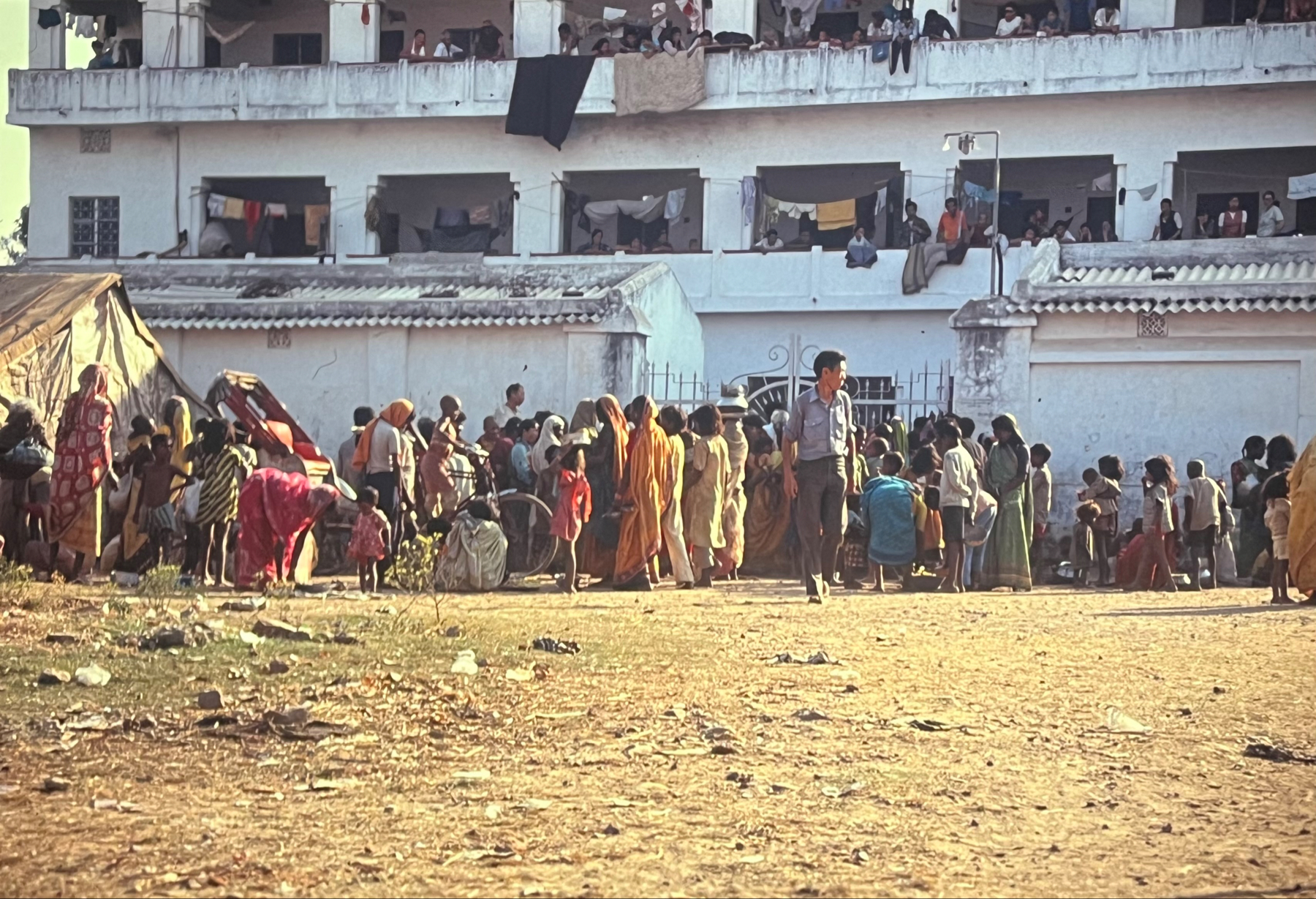 A crowd of people, many wearing traditional Indian clothing, gathered outside a white two-story building with balconies where more individuals are standing or sitting. Some are queuing toward a tent, suggesting a possible event or distribution taking place. The ground is dusty