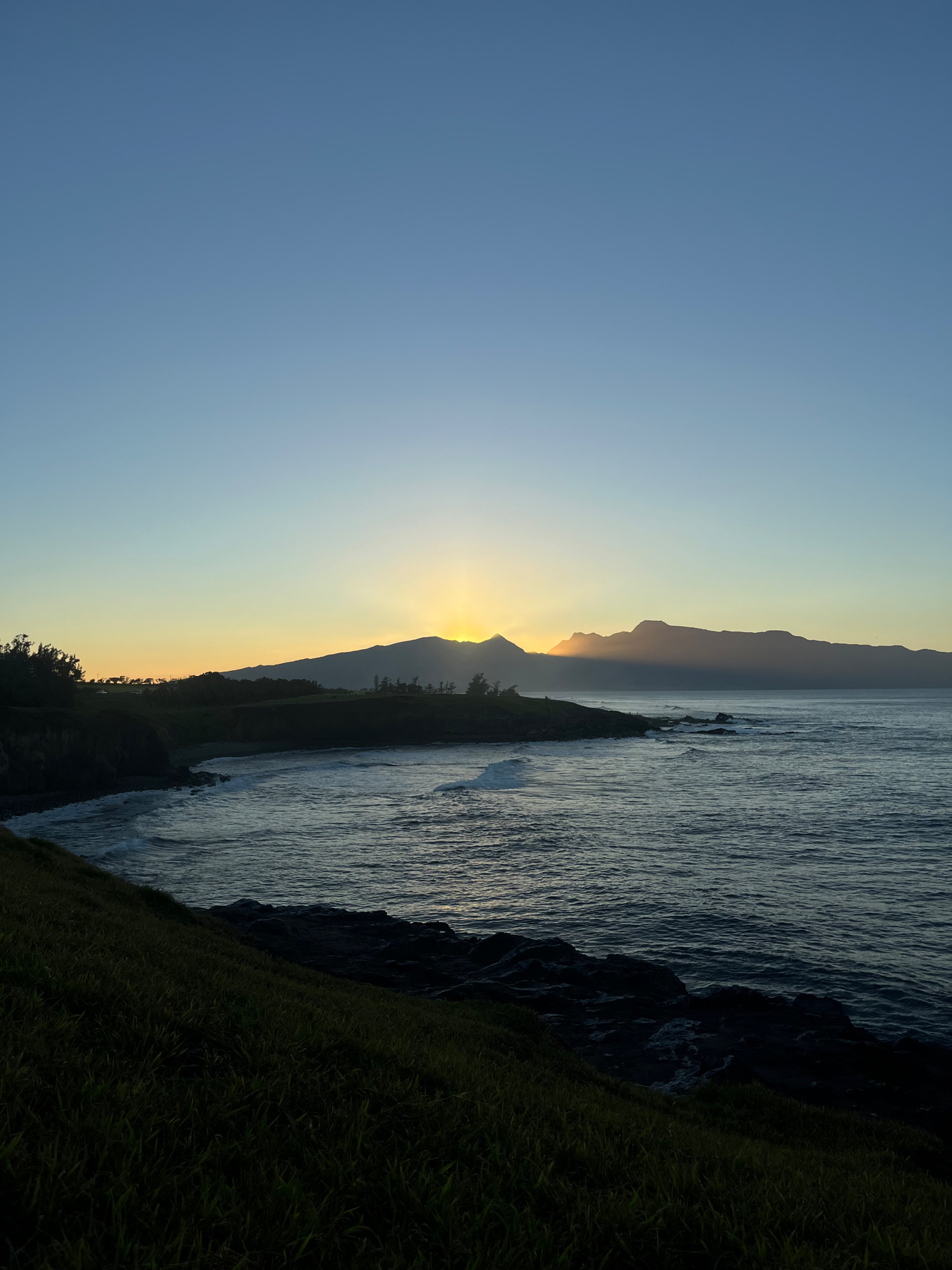 Sunset over the ocean with the sun peeking through the silhouette of the West Maui mountains, with a grassy foreground and a rocky shoreline.