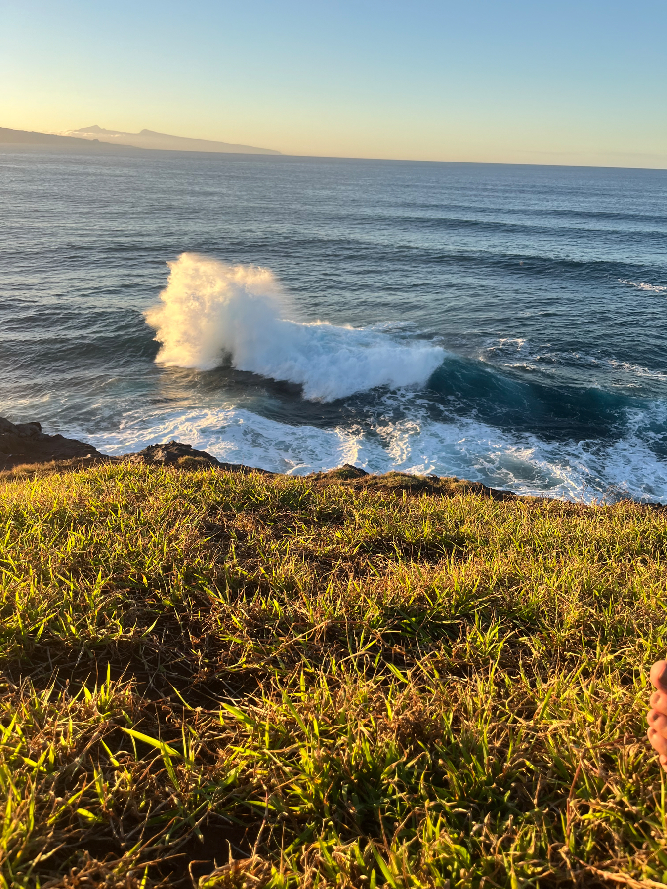 Ocean waves crashing against rocky coastline with grassy foreground and the Hawaiian island of Molokai in the distance at sunset.