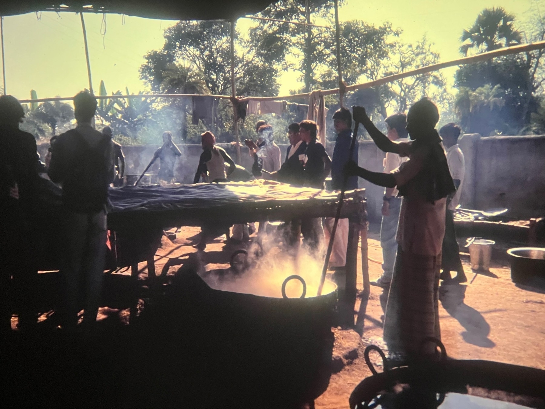 A hot cauldron being stirred while men stand around in a large area covered demarcated with a bamboo frame, some covered