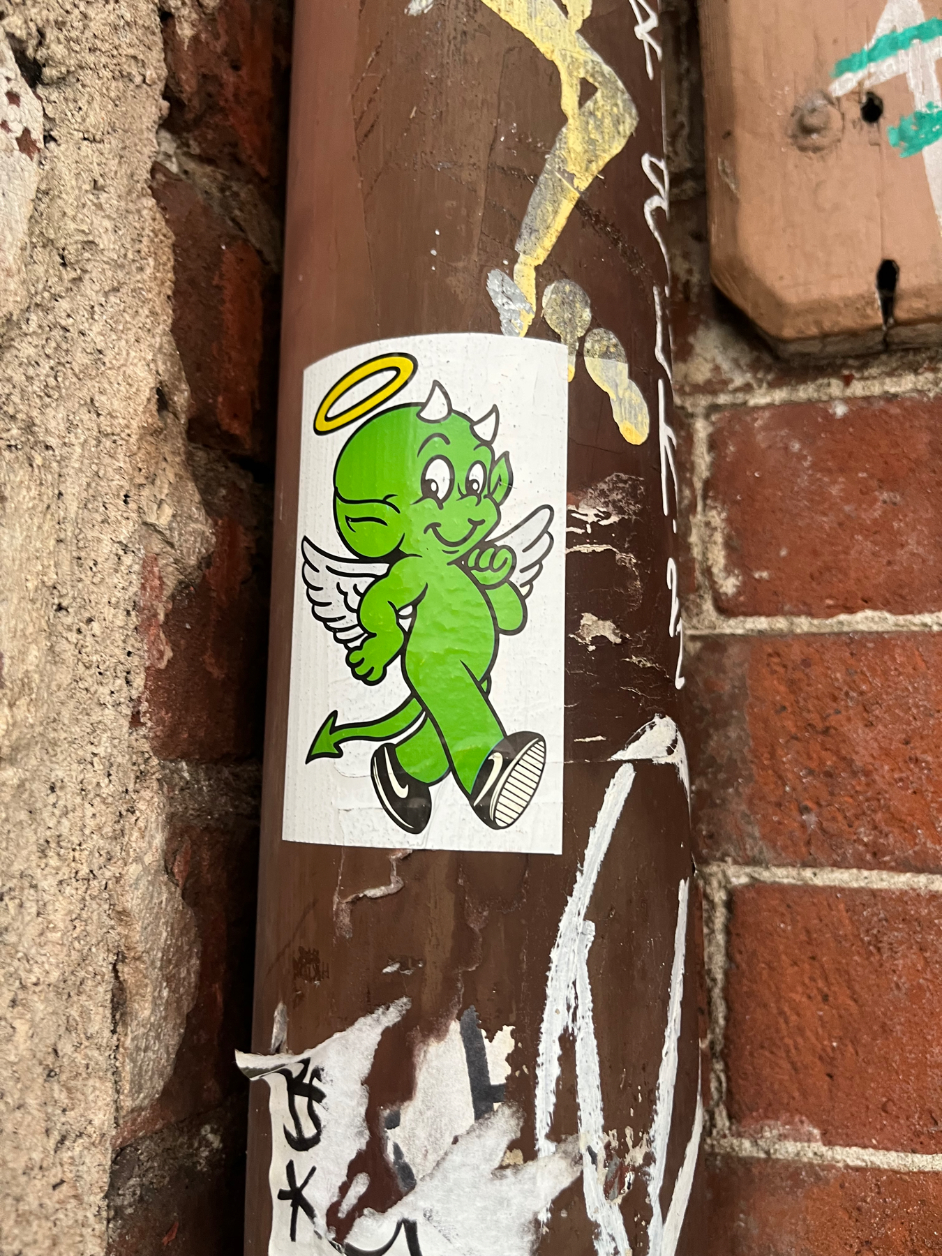 A sticker on a pipe depicting a walking green devil with black shoes on, small white wings, a pointy tail, a smiley face with horns on top, and above him a halo