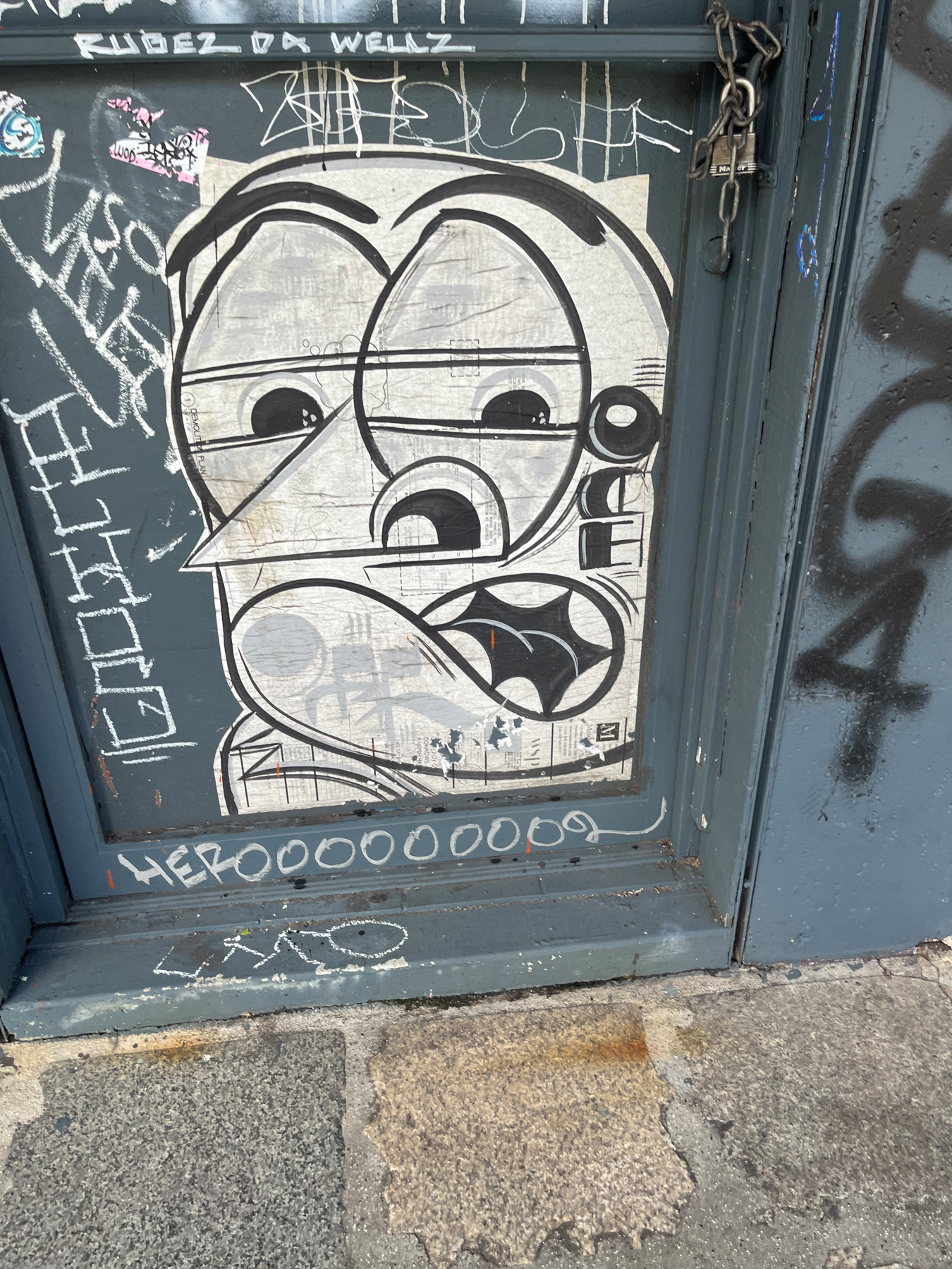 Graffiti on a doorway of a face, big eyes, triangular nose, with a concerned and worried expression. Various scribbles around and a padlock and chain hikding the door locked.