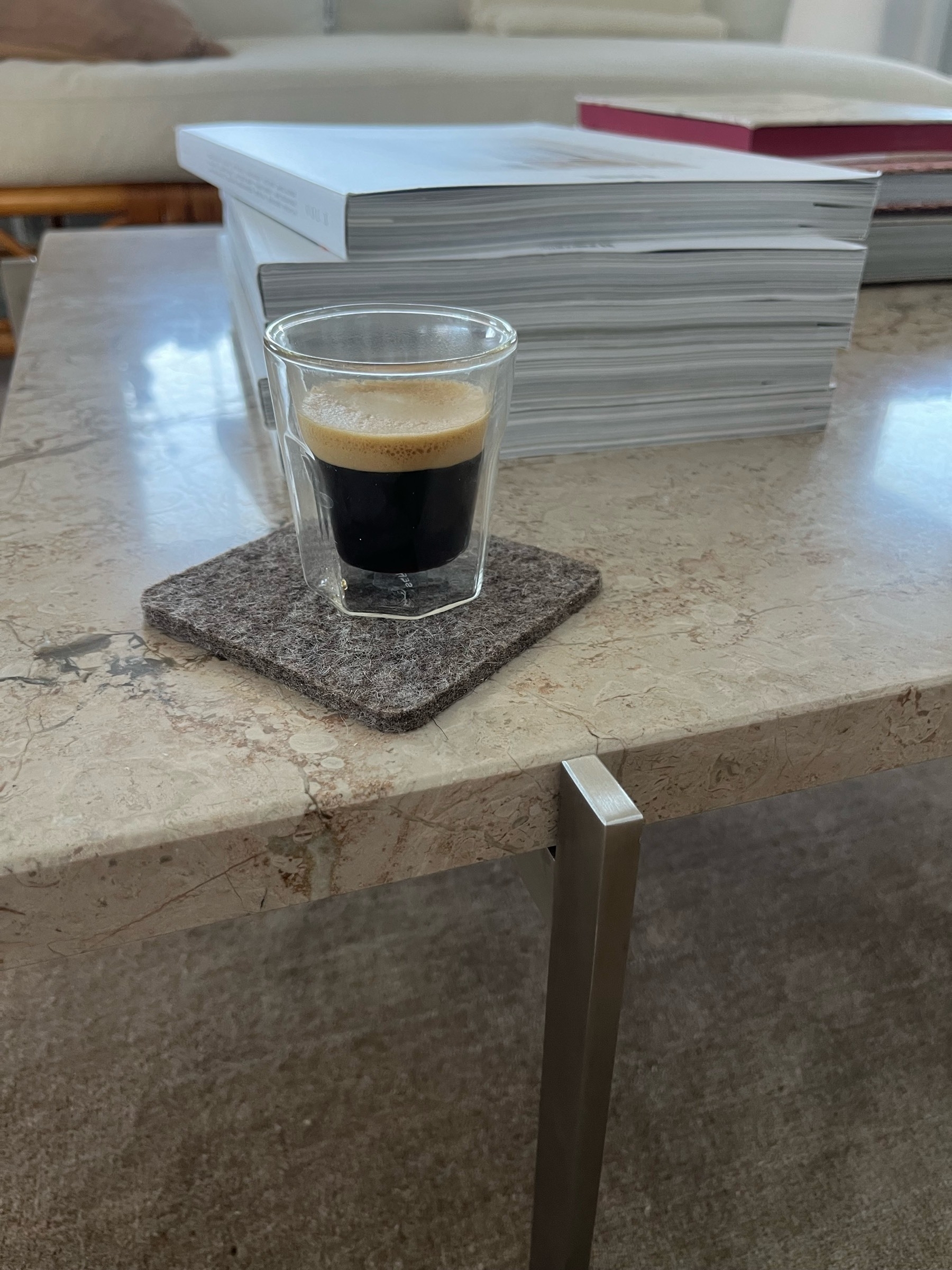 A cup of espresso coffee sitting on a mat on a marble table with a pile of white books behind it