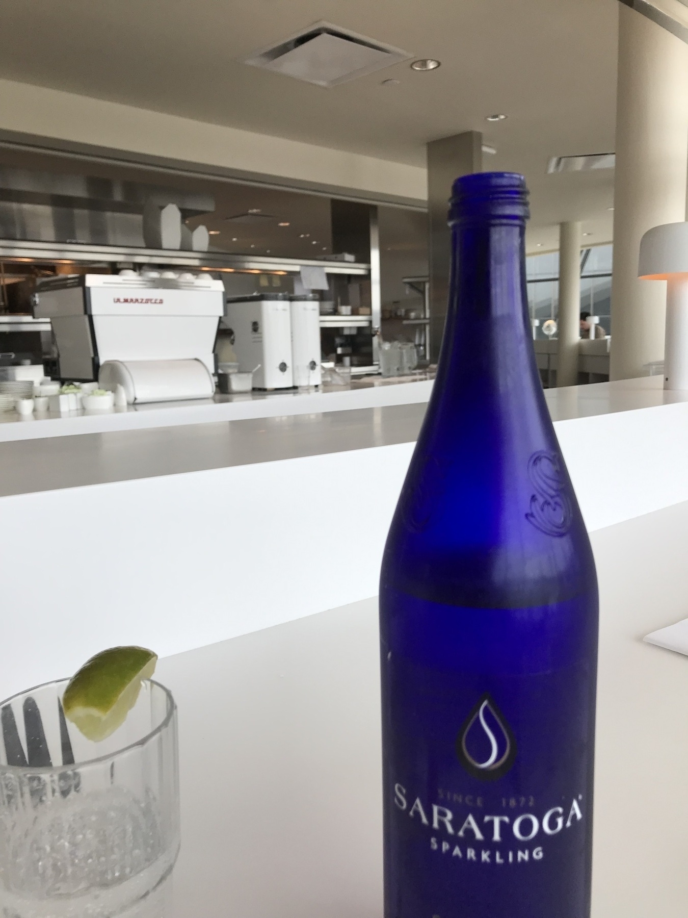A blue bottle of sparkling water alongside a glass of sparkling water. In the background is a restaurant kitchen