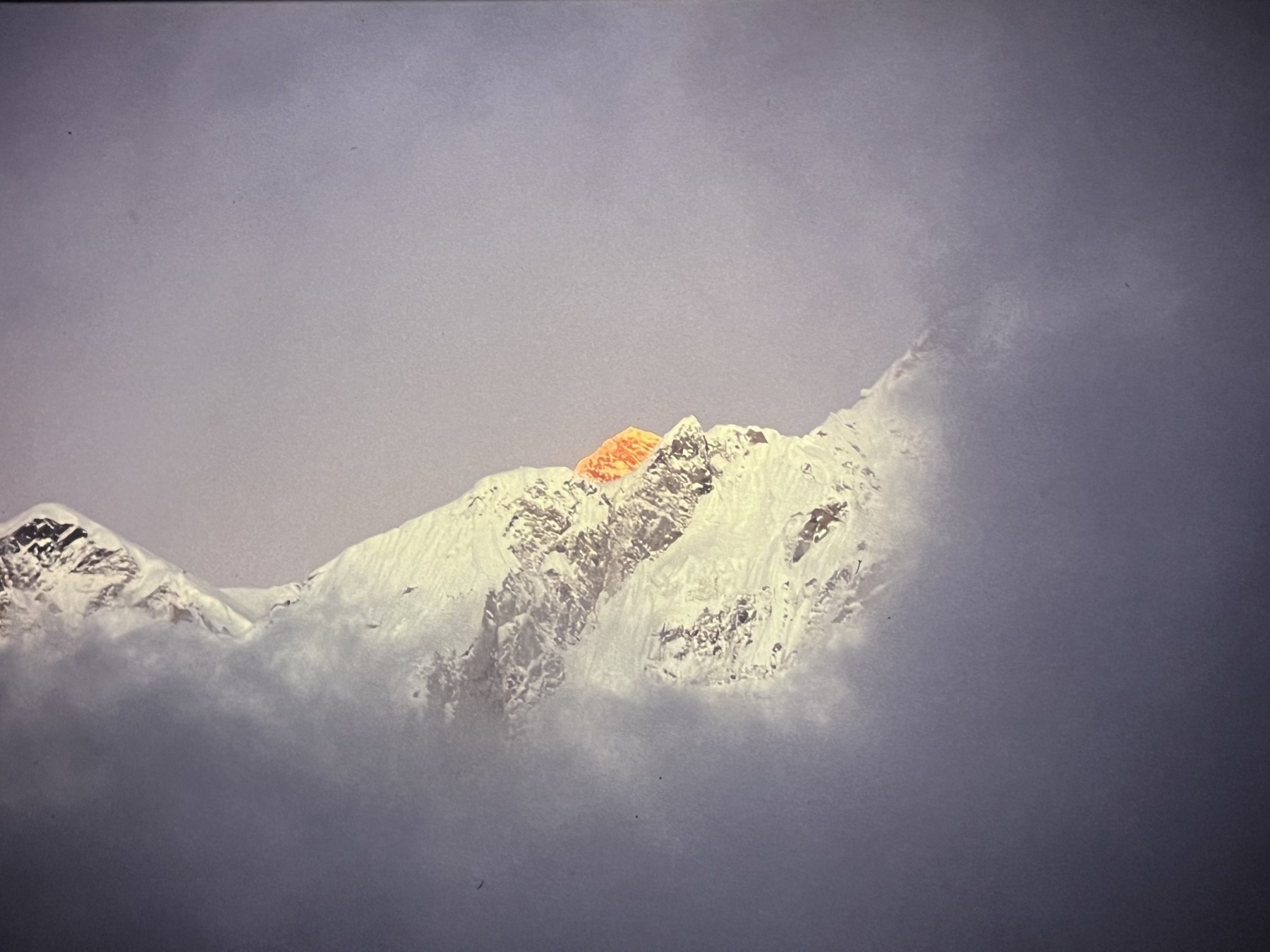 The summit of Mt Everest, peeking out from behind surrounding mountains, coloured orange as it catches the last rays of sunlight at the end of the day. Clouds are building up