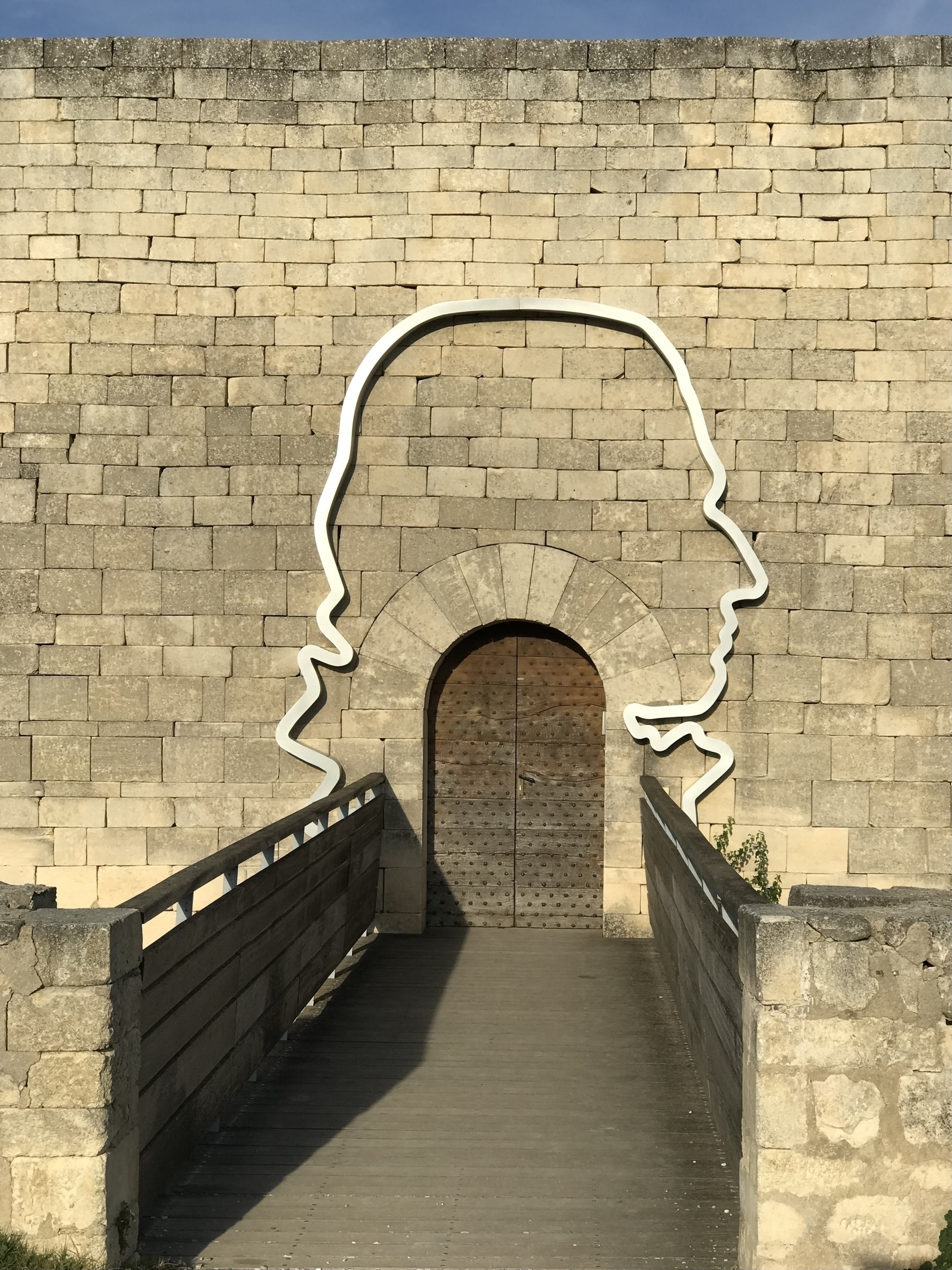 The outline of a person maybe Shakespeare, on a wall surrounding a door, approached by a wooden bridge.