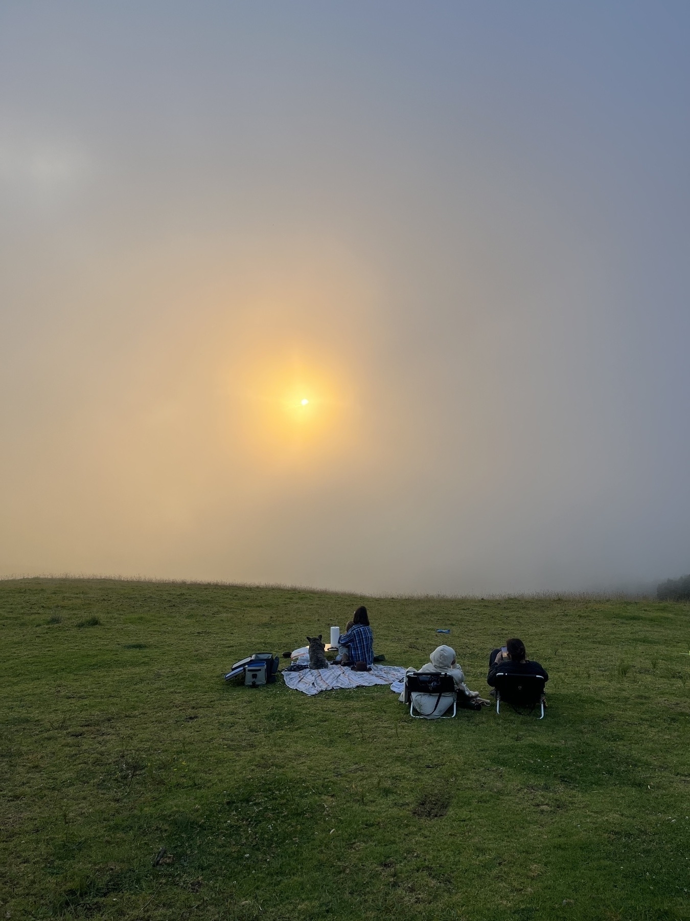 The sun pears through a thick cloud fog as people and a dog sit around a picnic blanket looking on.