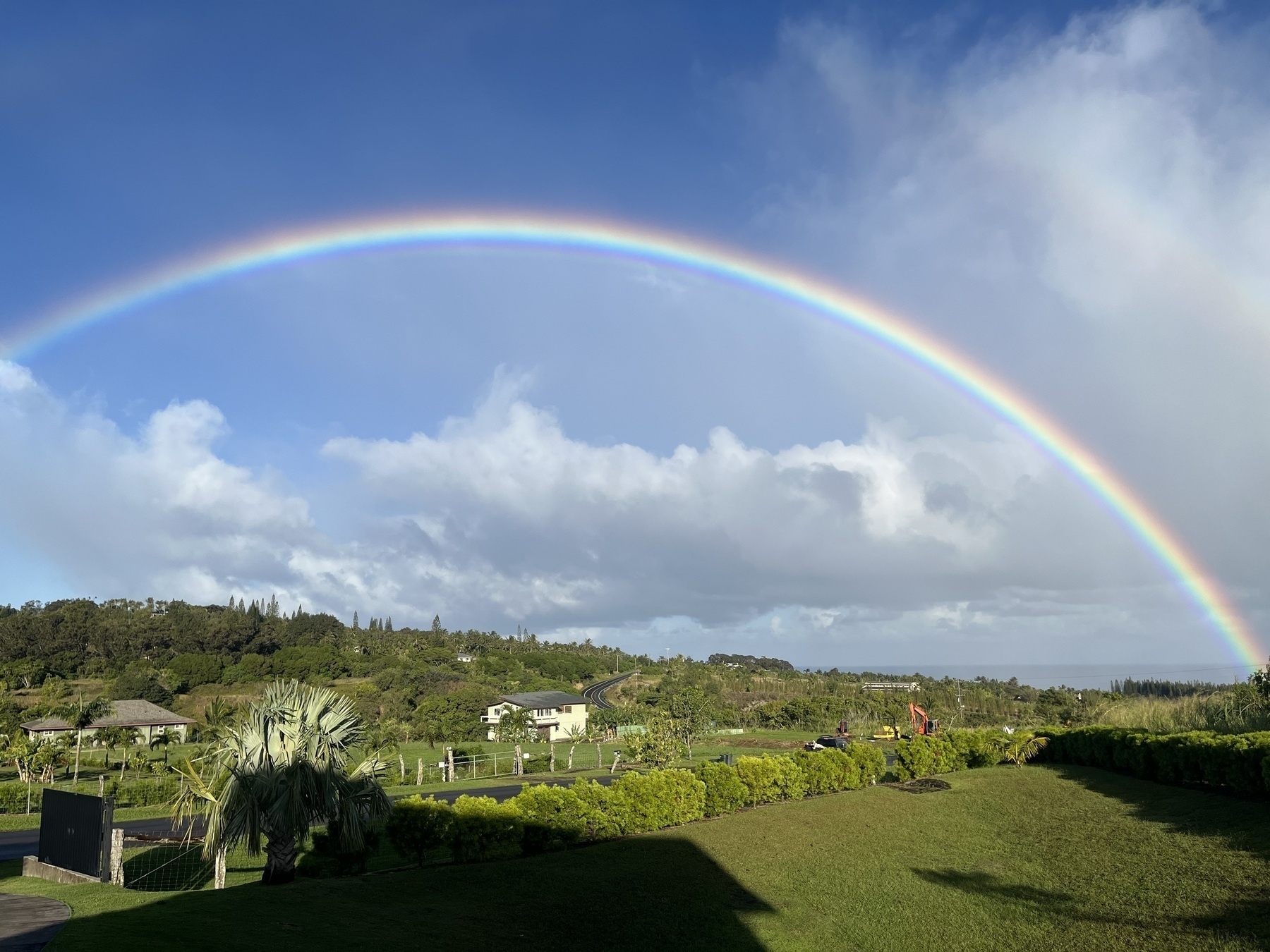A full rainbow arching across a blue sky with scattered cloud. Beneath the rainbow is a tropical rural scene with a couple of houses a road going up a hill and curving away. The grey ocean is in the distance