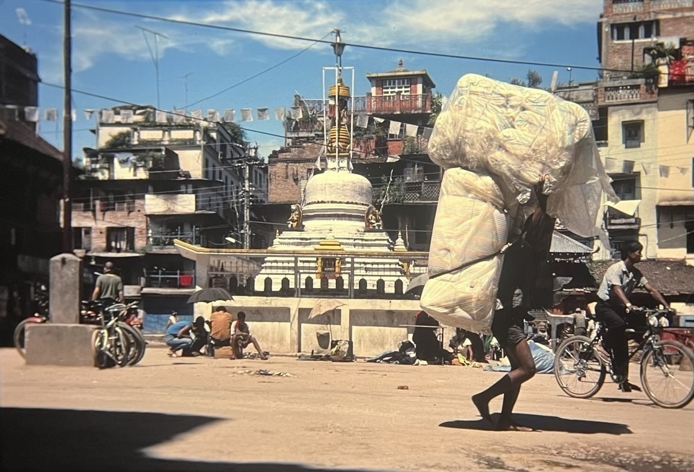 A small square in Kathmandu surrounded by many buildings. In the middle is an old Buddhist Stupa with prayer wheels around it. A string of old prayer flags hang from its top. In front a group of men sit on the ground, one with an umbrella to shade from the sun. A klutch of bicycles lie against a concrete base of a pole. To the right a man cycles away. In the foreground a porter carries an enormous white load using a hemp rope over his forehead