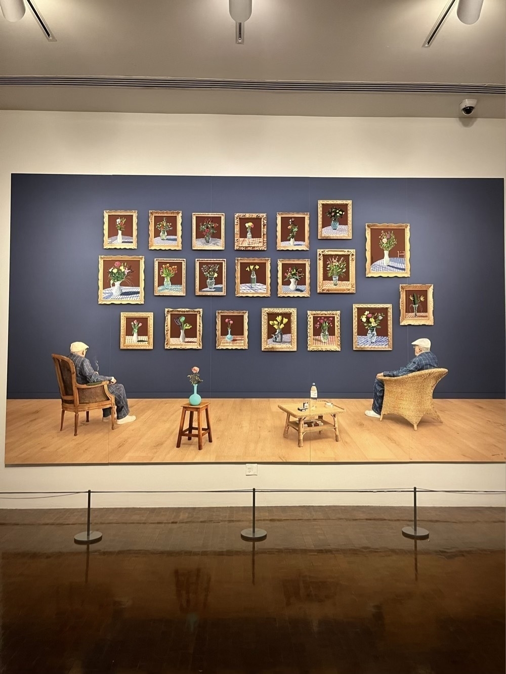 A huge David Hockney painting with two images of himself sat in chairs viewing prints that he has created.
