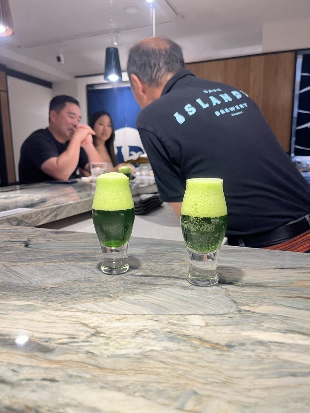 Two small glasses of green matcha beer, an IPA, with the server in the background serving two other customers.