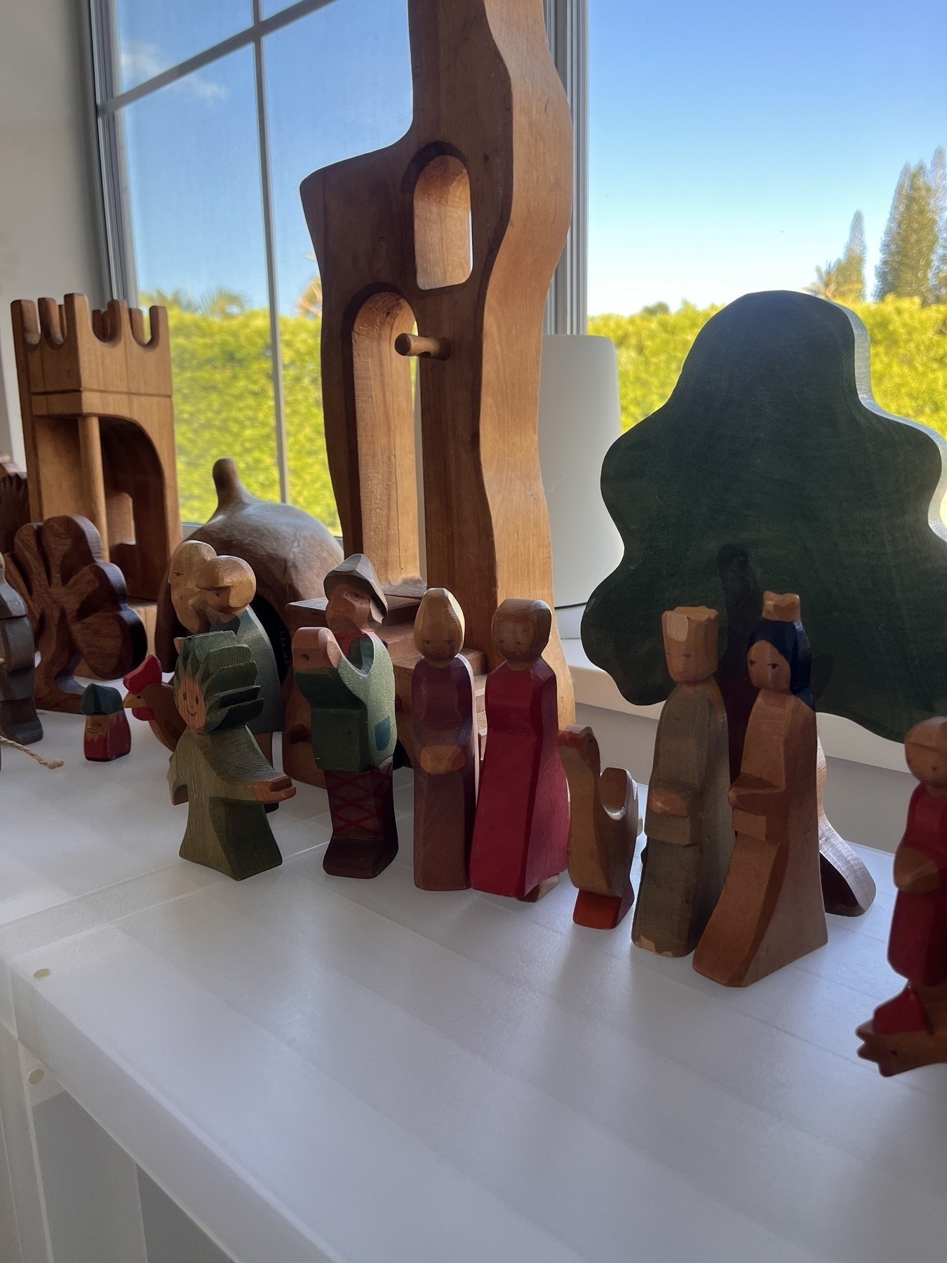 A row of wooden toys in the shape of castles, people and animals, all sitting on a shelf with a window behind. Outside there is blue sky and a green hedge.