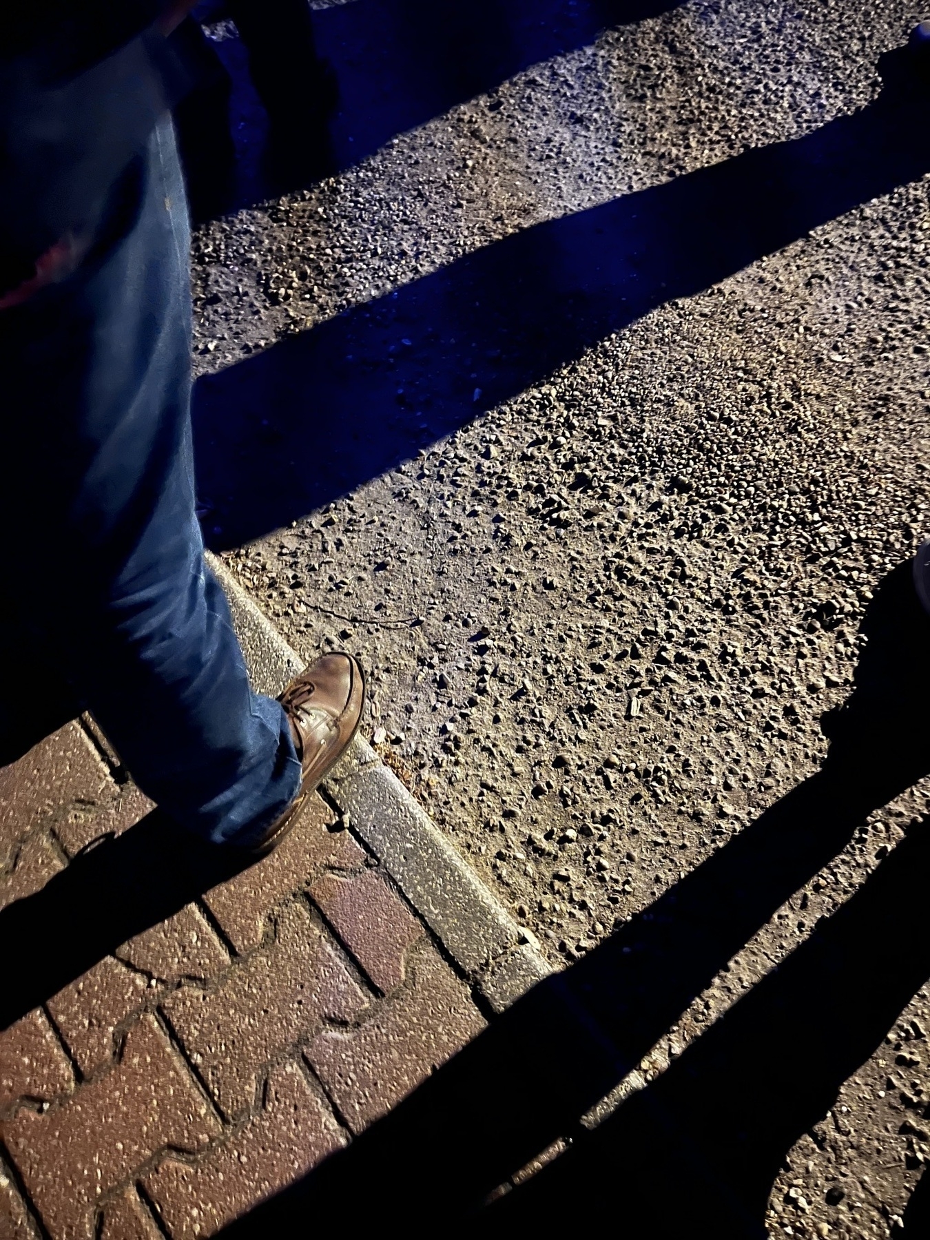 The leg and feet of someone standing outside at night and shadows cast by other people as the people listen to music at night.
