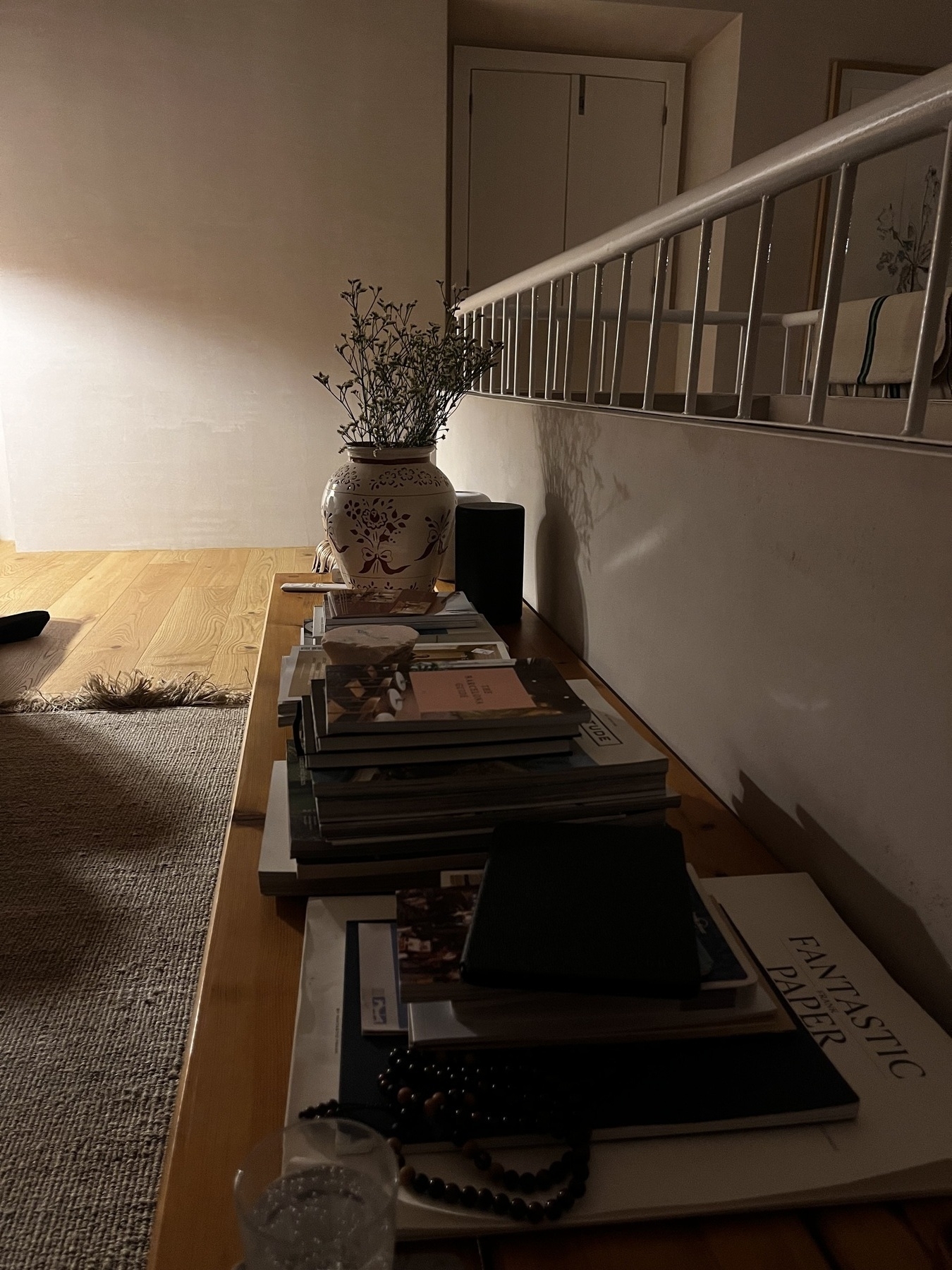 A nighttime scene in a house. A bench with books and a vase of flowers, alongside a low wall and railings. A shuttered window behind and light from the side.