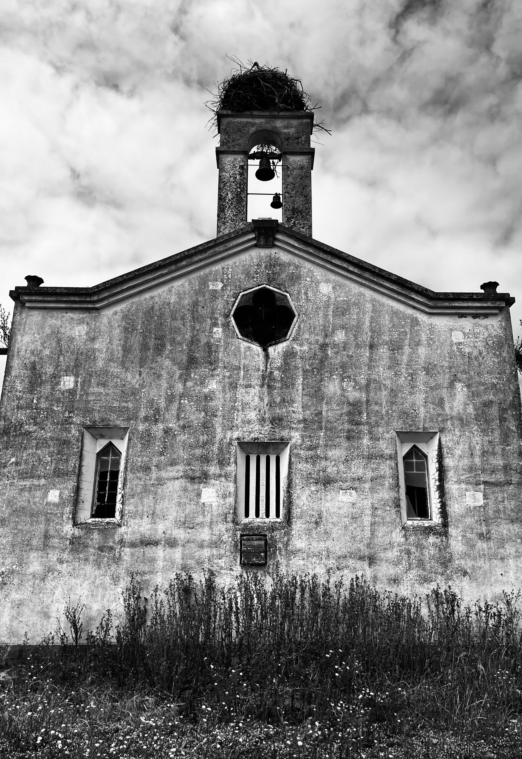 A black and white photograph of the end of an old church, with a bell on top and a stalk’s nest on top of that.