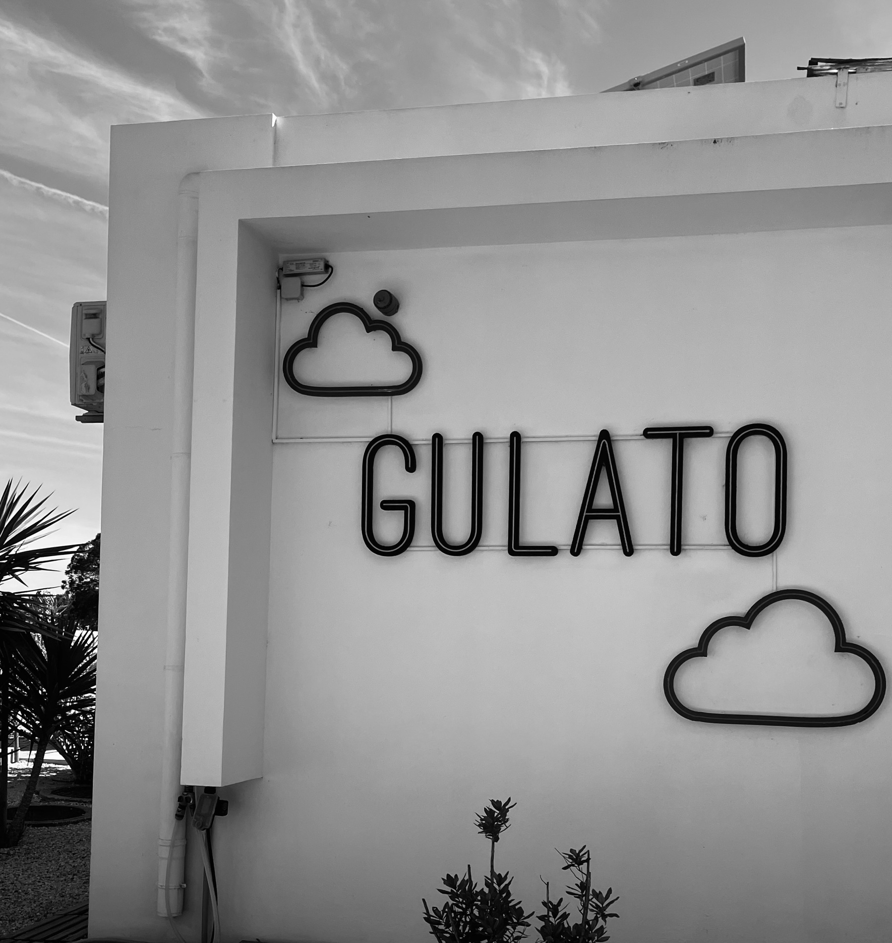 A black and white photo of the sign on a Gelato Store. The word Gulato with two clouds all on a white wall
